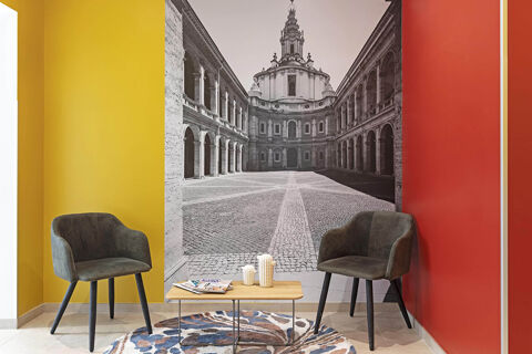 Interior corner of B&B Hotel Roma San Lorenzo Termini with yellow and red walls, two grey chairs, a wooden table, and a black and white mural