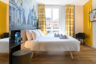 Minimalist hotel room with white bed linens, yellow patterned curtains, black and white wall art, and wooden floor at B&B Hotel Roma San Lorenzo Termini