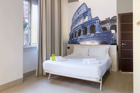 Room at B&B Hotel Roma Trastevere with a bed, white linens, folded towels, a large wall photo of the Colosseum, and a window