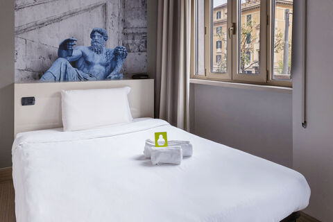 A minimalist hotel room at B&B Hotel Roma Trastevere with a white bed, folded towels, a pillow, and wall art of a reclining statue