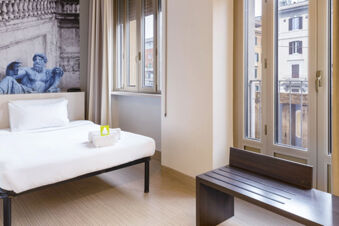 Minimalist hotel room with a bed, white linens, towel set, large windows, bench, and statue wallpaper at B&B Hotel Roma Trastevere