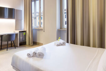 A hotel room at B&B Hotel Roma Trastevere with a neatly made bed, rolled towels, a desk with a chair, TV, and two windows with curtains