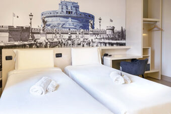 Two single beds with white linens and rolled towels in a room with a desk, chair, and B&B Hotel Roma Trastevere mural wall art