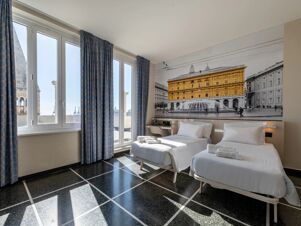 Sunlit room at B&B Hotel Genova Principe with two single beds, blue patterned curtains, and large wall art of a historic building