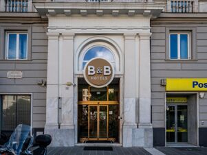 Entrance of B&B Hotel Genova Principe with a circular brown and white sign above glass doors on a marble and stone building facade