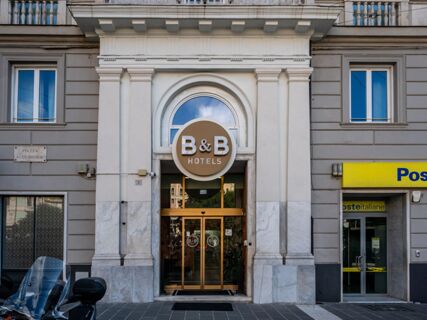 Entrance of B&B Hotel Genova Principe with a circular brown and white sign above glass doors on a marble and stone building facade