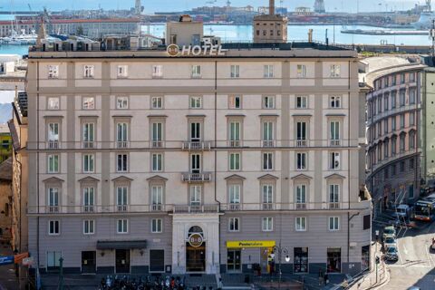 A large beige building with multiple windows and the B&B Hotel Genova Principe sign on top near a busy street and waterfront harbor background