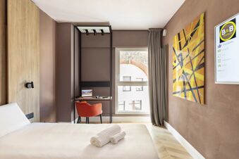 Hotel room at B&B Hotel Genova City Center with a white bed, rolled towels, a red chair, desk, window, and abstract wall art