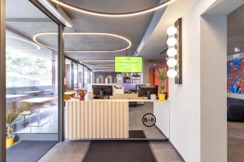 Reception area with wooden front desk, two computer monitors, modern circular ceiling lights, and a glass wall at B&B Hotel Genova City Center