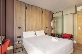 Bed with white pillows and linens in a room with wooden wall panels, red chair, and luggage stand at B&B Hotel Genova City Center