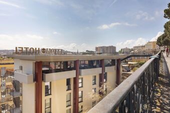 B&B Hotel Genova City Center rooftop sign with beige building, street railing, trees, and cityscape under partly cloudy sky