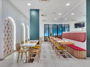 Dining area with wooden tables, mixed chairs, patterned arched windows, red cushioned seating, and a TV screen at B&B Hotel Bergamo City