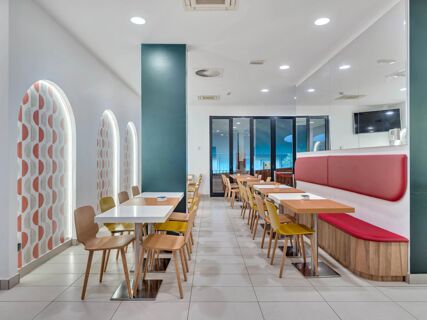 Dining area with wooden tables, mixed chairs, patterned arched windows, red cushioned seating, and a TV screen at B&B Hotel Bergamo City