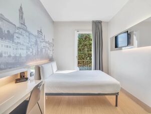 Modern hotel room at B&B Hotel Bergamo City with a white bed, wall art, desk with chair, window, and wall-mounted TV