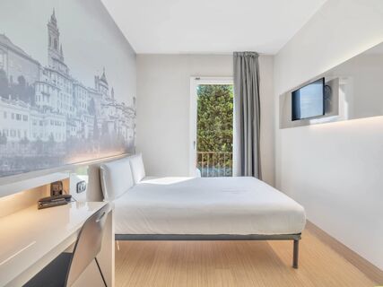 Modern hotel room at B&B Hotel Bergamo City with a white bed, wall art, desk with chair, window, and wall-mounted TV