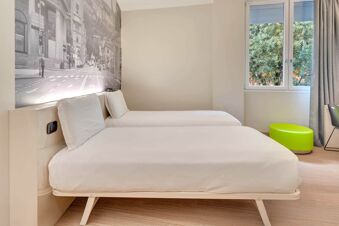Two single beds with white linens and pillows are placed against a wall with a black and white cityscape mural at B&B Hotel Bergamo City
