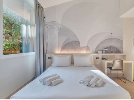 Double bed with white pillows and towels folded on it, window with gray curtains, desk with chair in B&B Hotel Bergamo City room