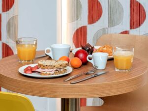 Breakfast table at B&B Hotel Bergamo City with two glasses of orange juice, white cups, fresh fruit, croissants, and sandwich pieces