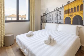 Bright hotel room at B&B Hotel Brescia with white striped bed, rolled towels, yellow patterned curtains, and a cityscape mural