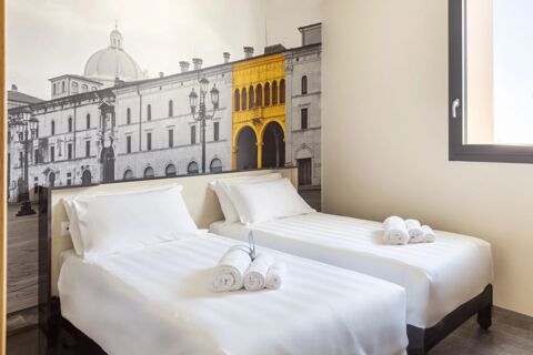Two single beds with white linens and rolled towels face a large black and white photo wall with a yellow accent in a B&B Hotel Brescia room