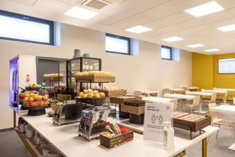 Breakfast buffet with bread rolls, apples, packaged snacks, coffee machines, and seating area at B&B Hotel Brescia