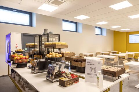 Breakfast buffet with bread rolls, apples, packaged snacks, coffee machines, and seating area at B&B Hotel Brescia