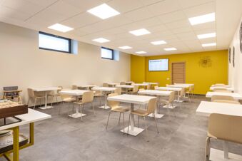 Minimalist dining room with white square tables, beige chairs, a yellow accent wall, and a screen at B&B Hotel Brescia