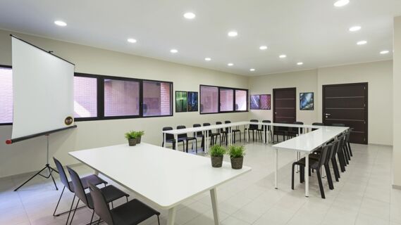 Meeting room with U-shaped white tables, black chairs, framed pictures, projector screen, and plants at B&B Hotel Como Camerlata