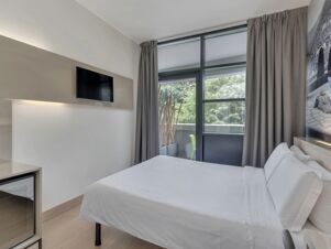 A modern hotel room at B&B Hotel Como Camerlata with a large window covered by gray curtains and a mounted flat-screen TV