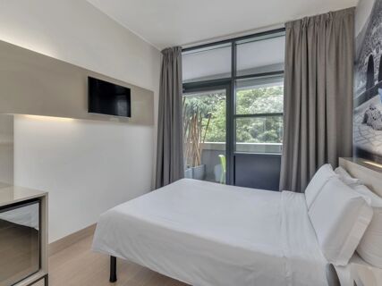 A modern hotel room at B&B Hotel Como Camerlata with a large window covered by gray curtains and a mounted flat-screen TV