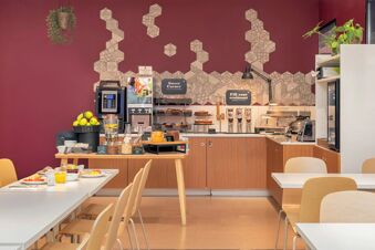 Breakfast area with wooden chairs and tables, cereal dispensers, juice machines, and decorated wall at B&B Hotel Como Camerlata