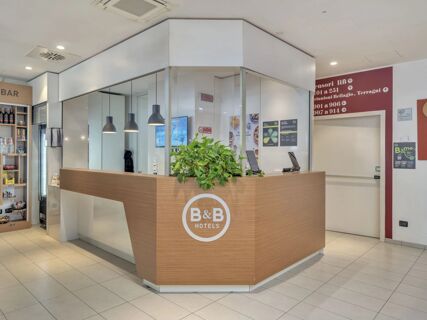 Reception area of B&B Hotel Como Camerlata with a wooden front desk, green plant, hanging lights, and a snack bar visible to the side