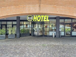 The entrance of B&B Hotel Como Camerlata features a curved yellow sign above a revolving glass door with potted plants nearby