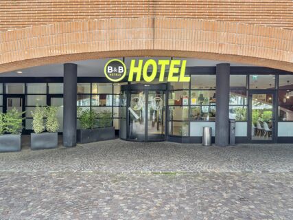 The entrance of B&B Hotel Como Camerlata features a curved yellow sign above a revolving glass door with potted plants nearby