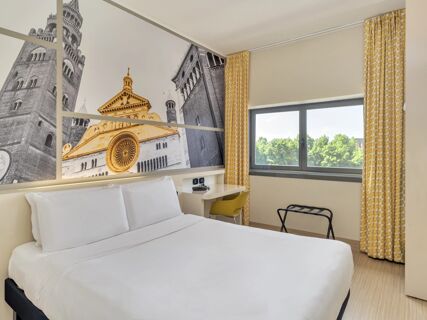 Bed with white linens, yellow patterned curtains, desk with yellow chair, and large wall artwork in B&B Hotel Cremona room