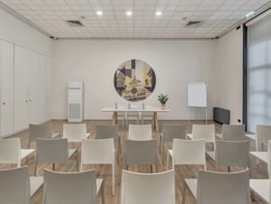 Conference room setup at B&B Hotel Cremona with rows of white chairs, a table with water bottles, and wall art of buildings