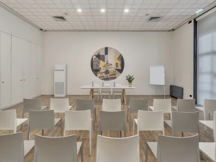 Conference room setup at B&B Hotel Cremona with rows of white chairs, a table with water bottles, and wall art of buildings