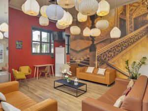 Modern lounge area at B&B Hotel Cremona with orange couches, yellow chairs, hanging paper lanterns, and a decorative wall mural of stairs