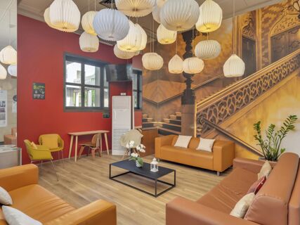 Modern lounge area at B&B Hotel Cremona with orange couches, yellow chairs, hanging paper lanterns, and a decorative wall mural of stairs