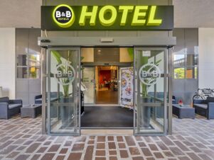 Entrance of B&B Hotel Cremona with glass doors open, potted plants inside, outdoor seating, and a large yellow hotel sign above