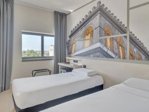 Hotel room at B&B Hotel Cremona with two single beds, a window with gray curtains, a desk, and a large wall photo of a historic building with golden windows