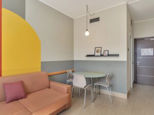 Interior of B&B Hotel Cremona with a brown sofa, four white chairs around a round table, wall art, and a hanging light bulb
