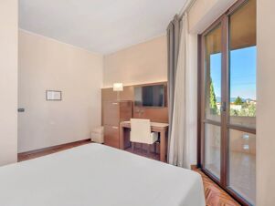 A room at B&B Hotel Habitat Giussano featuring a bed, wooden desk with chair, wall-mounted TV, floor lamp, and large window with curtains