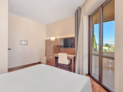 A room at B&B Hotel Habitat Giussano featuring a bed, wooden desk with chair, wall-mounted TV, floor lamp, and large window with curtains
