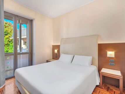Bedroom at B&B Hotel Habitat Giussano with a large bed, white linens, two pillows, wooden nightstands, and a glass door window