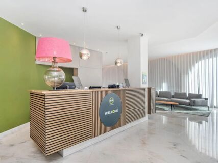 Reception desk with wood slats and a pink lamp in front of a green wall at B&B Hotel Habitat Giussano lobby area with seating