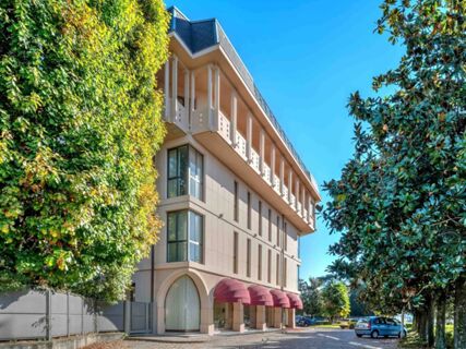 Modern beige building with large windows and red awnings surrounded by green trees under a blue sky at B&B Hotel Habitat Giussano