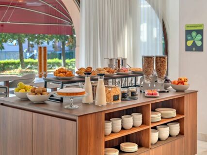 Breakfast buffet with cereals, milk, fruit, pastries, muffins, and plates on a wooden counter at B&B Hotel Habitat Giussano