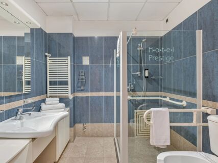 Bathroom with blue and beige tiles, white sink, towel rack, shower with glass door, and B&B Hotel Hotel Prestige Ornago sign