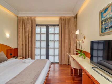 Bed with beige pillows and towels, patterned curtains, wall lamp, table with orchid, and TV in B&B Hotel Hotel Prestige Ornago